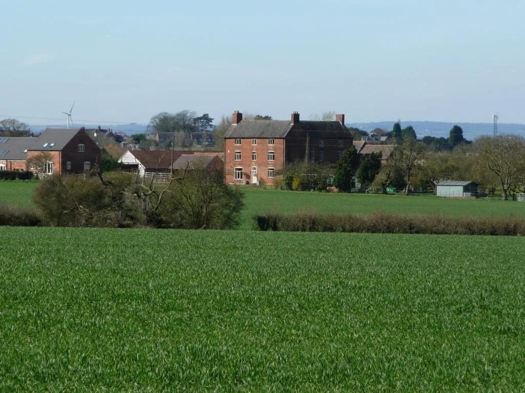 The three-storey brick Arkall farmhouse and outbuildings seen across a green wheat field.