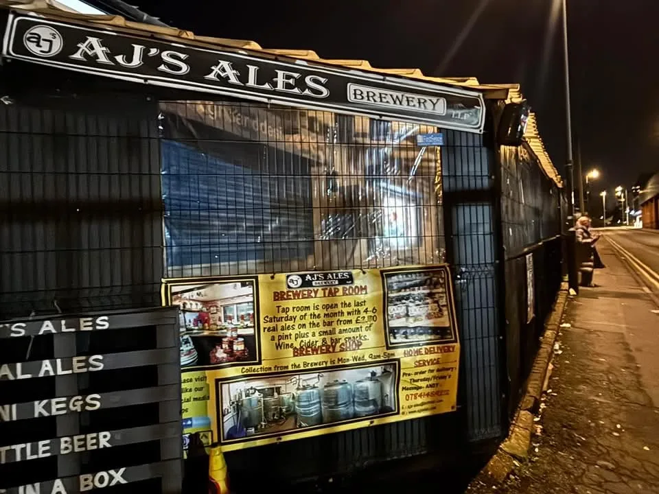 The AJ's Ales brewery sign and tap room notices on a fence beside a street at night