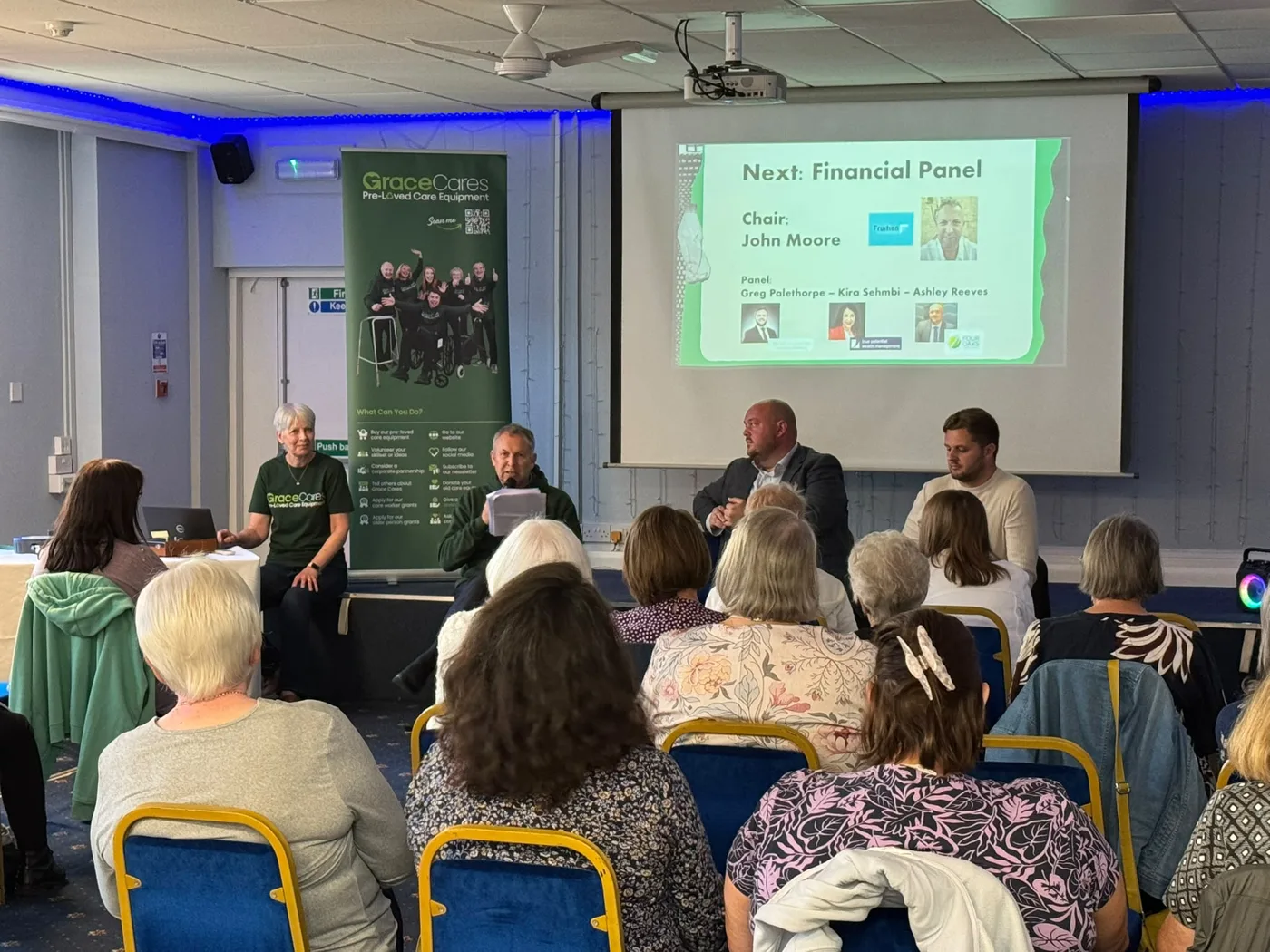 Carers seated in rows facing the financial panel at the front of a room at The Cathedral Hotel, with a Grace Cares banner and a screen reading Next: Financial Panel
