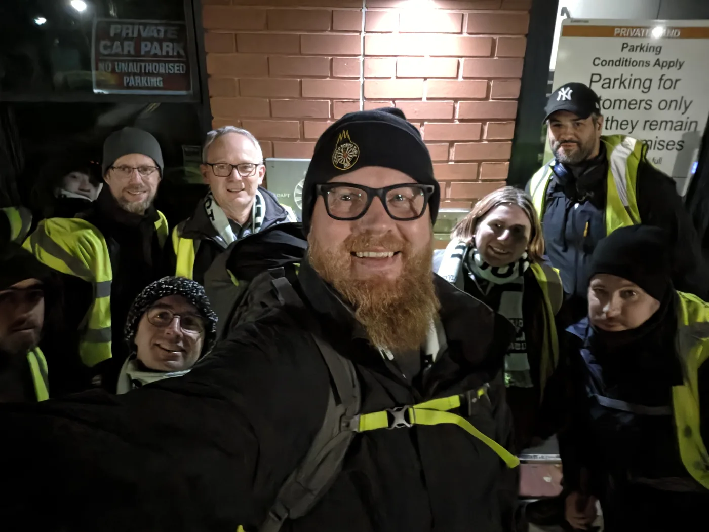 A group of walkers in hats and high-visibility vests posing for a selfie at night outside a brick building before the Lost Hours Walk