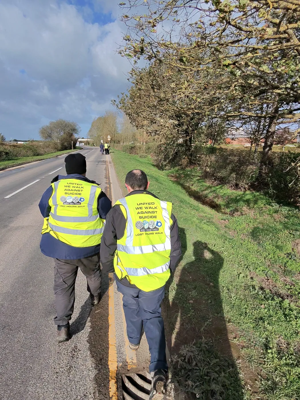 Two walkers seen from behind on a grass verge beside a country road, their yellow vests printed United We Walk Against Suicide, Lost Hours Walk