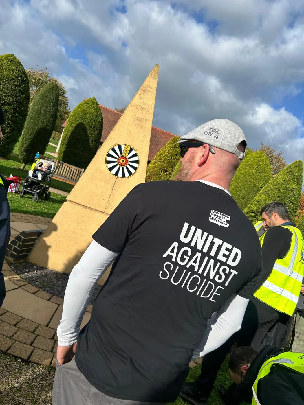 A walker seen from behind wearing a black CALM T-shirt printed United Against Suicide, beside a stone obelisk carrying the Round Table emblem