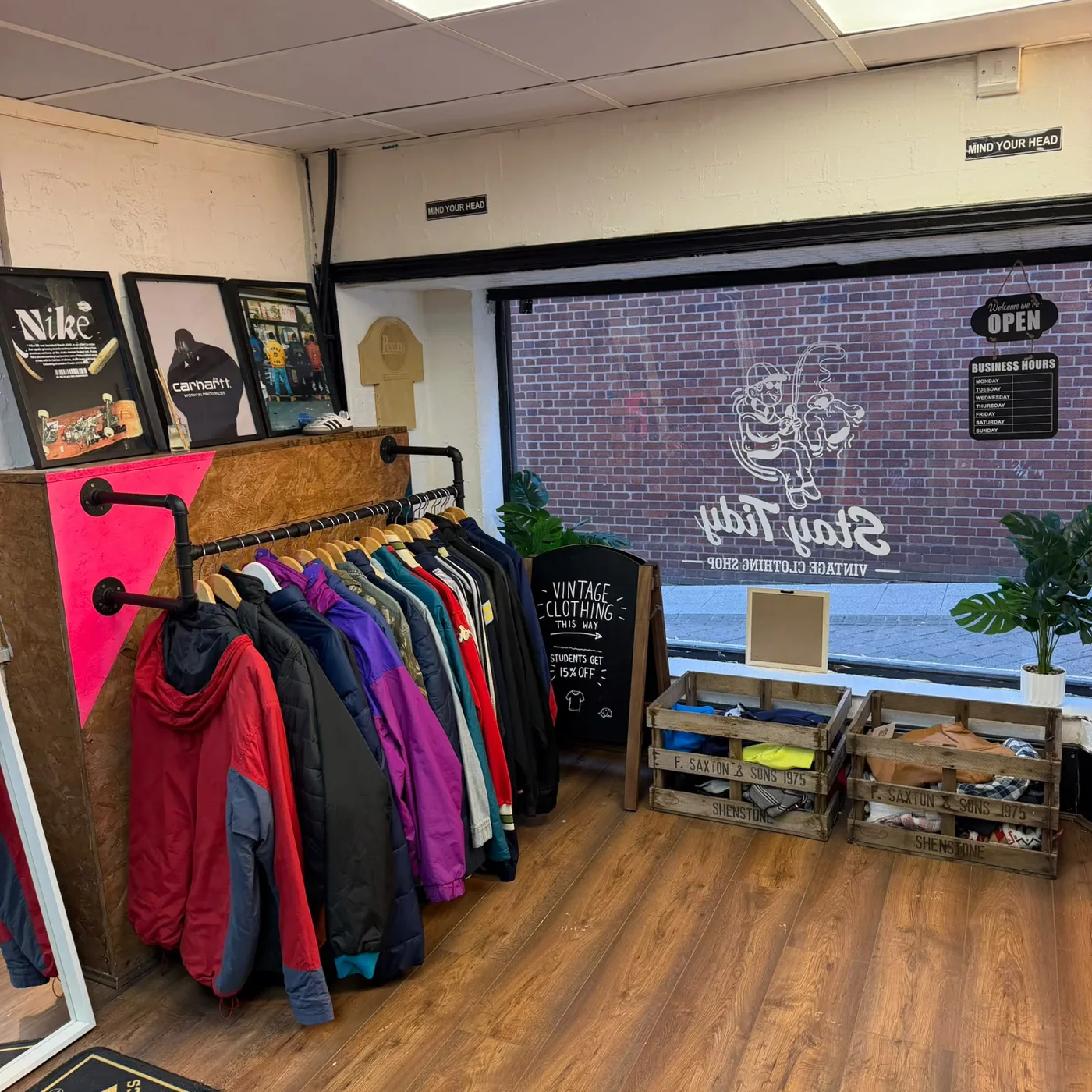 Inside Stay Tidy: a rail of colourful vintage jackets under framed Nike and Carhartt posters, a chalkboard reading Vintage clothing this way, students get 15% off, and two wooden crates of folded clothes by the window