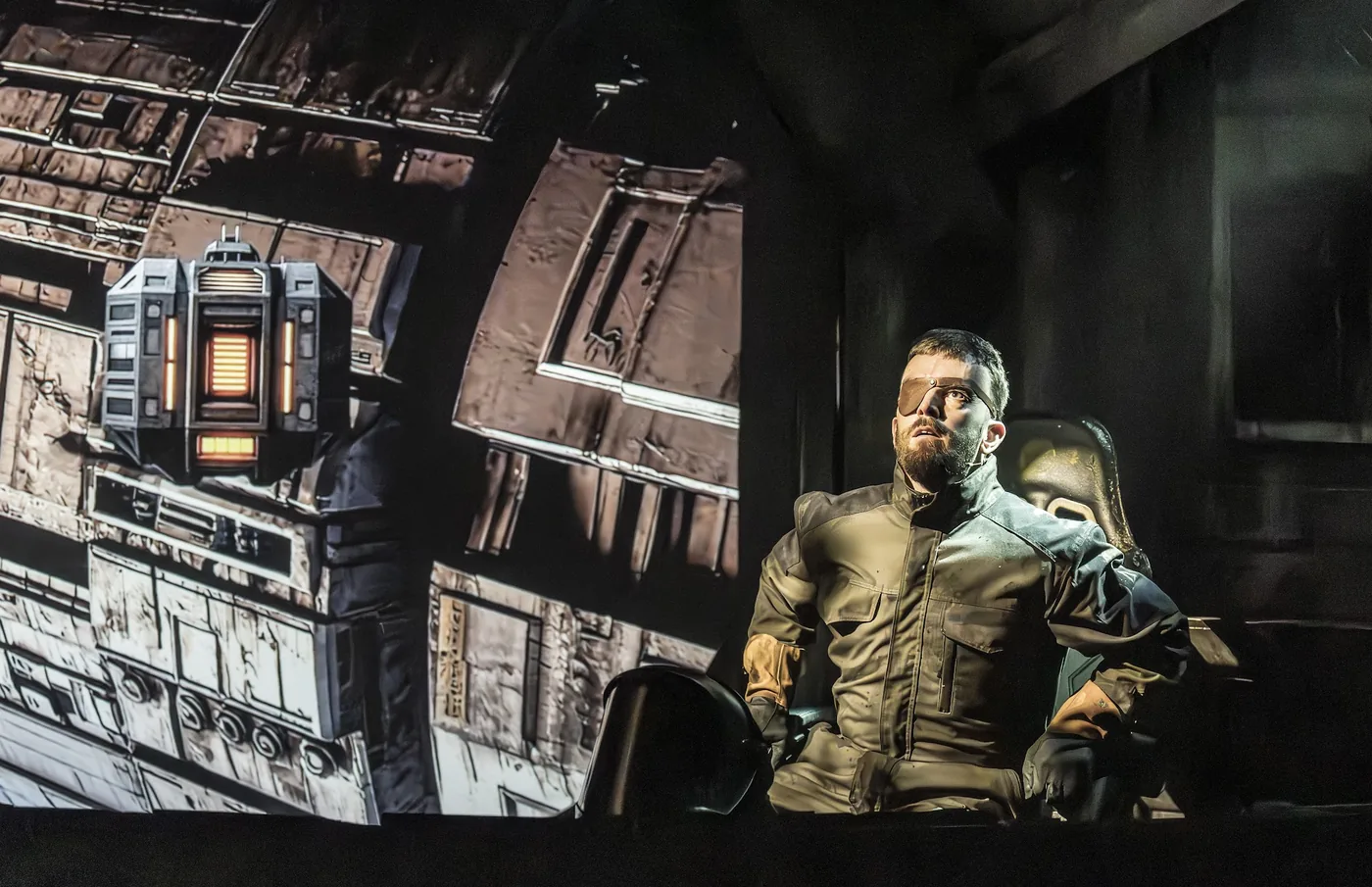 A performer in a flight suit looking up, with the hull of a spacecraft projected on the wall behind him