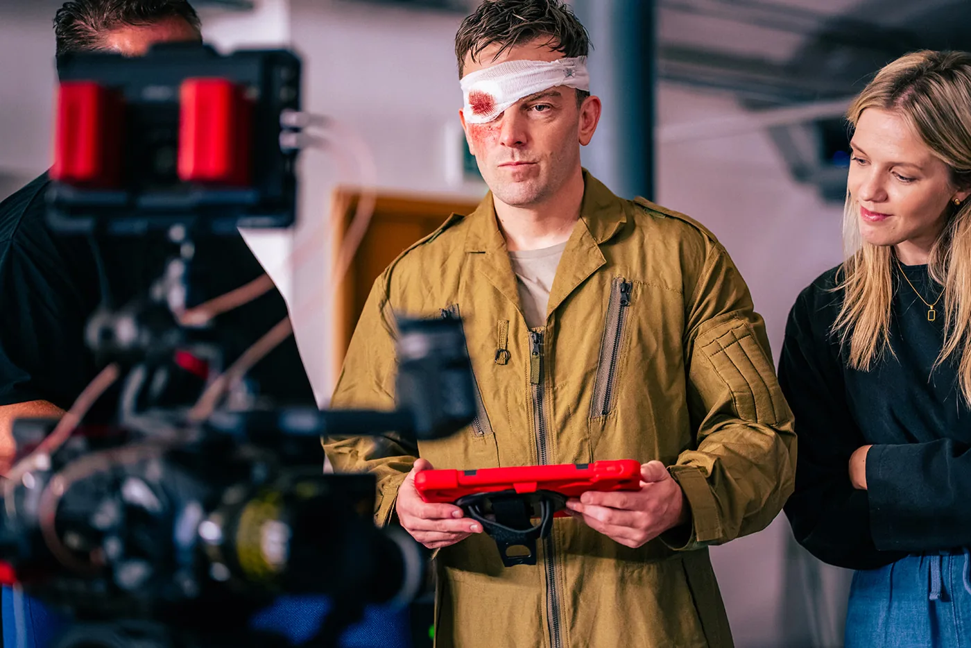 A film camera on a rig trained on a performer in a flight suit with a bloodied bandage over one eye, two crew members beside him